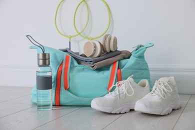 Photo of Stylish sports bag with sportswear, sneakers, water bottle, headphones and badminton rackets on floor near white wall indoors