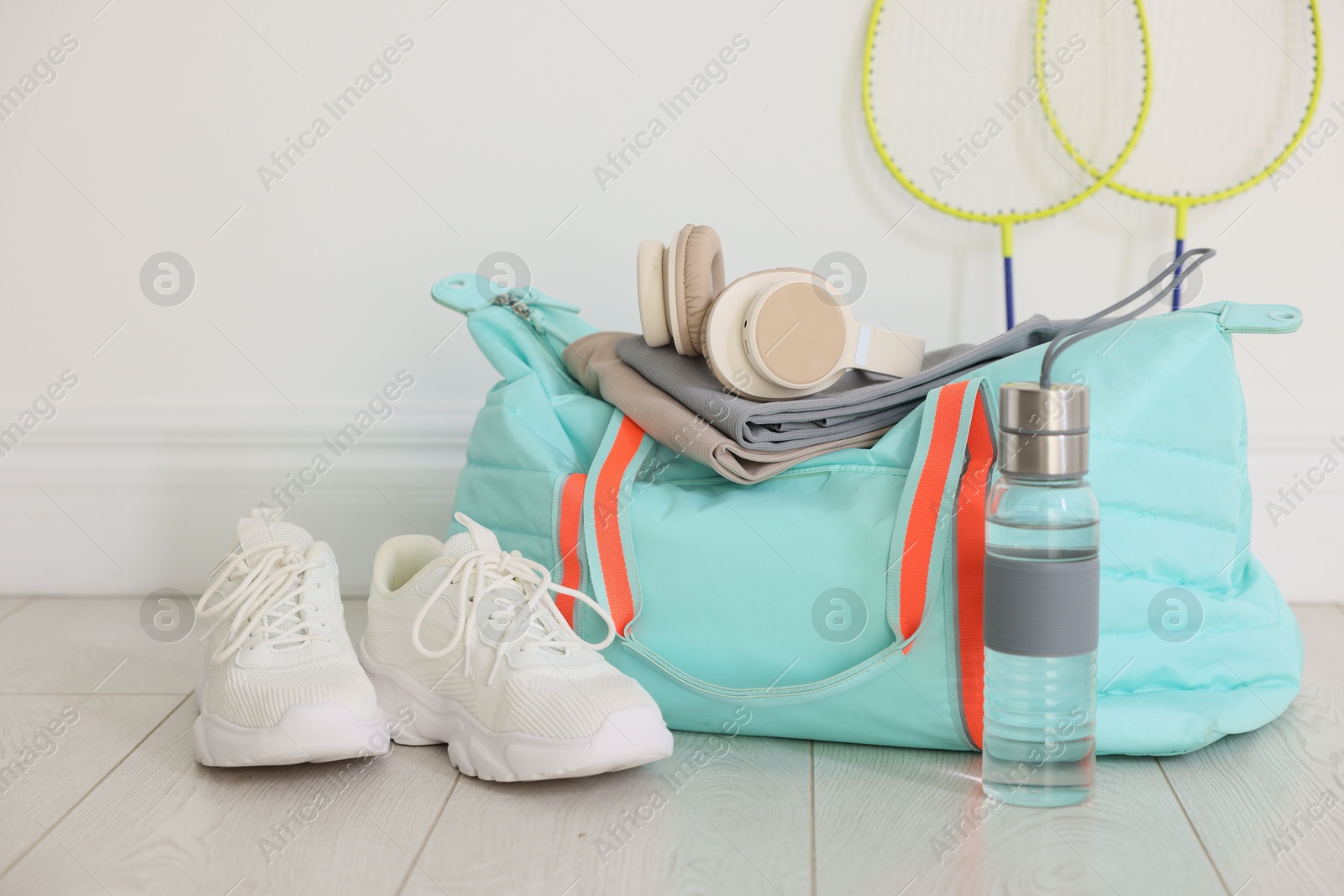 Photo of Stylish sports bag with sportswear, sneakers, water bottle, headphones and badminton rackets on floor near white wall indoors