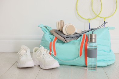 Photo of Stylish sports bag with sportswear, sneakers, water bottle, headphones and badminton rackets on floor near white wall indoors