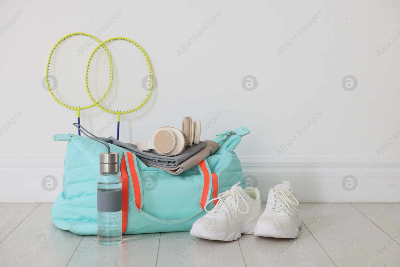 Photo of Stylish sports bag with sportswear, sneakers, water bottle, headphones and badminton rackets on floor near white wall indoors