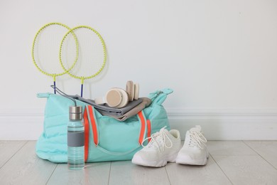 Photo of Stylish sports bag with sportswear, sneakers, water bottle, headphones and badminton rackets on floor near white wall indoors