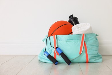 Photo of Stylish sports bag with basketball ball, skipping rope, water bottle and towel on floor near white wall indoors, space for text