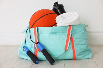 Photo of Stylish sports bag with basketball ball, skipping rope, water bottle and towel on floor near white wall indoors