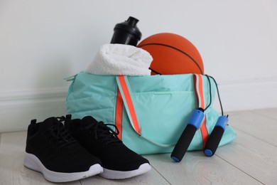 Photo of Stylish sports bag with basketball ball, skipping rope, water bottle, towel and sneakers on floor near white wall indoors, closeup