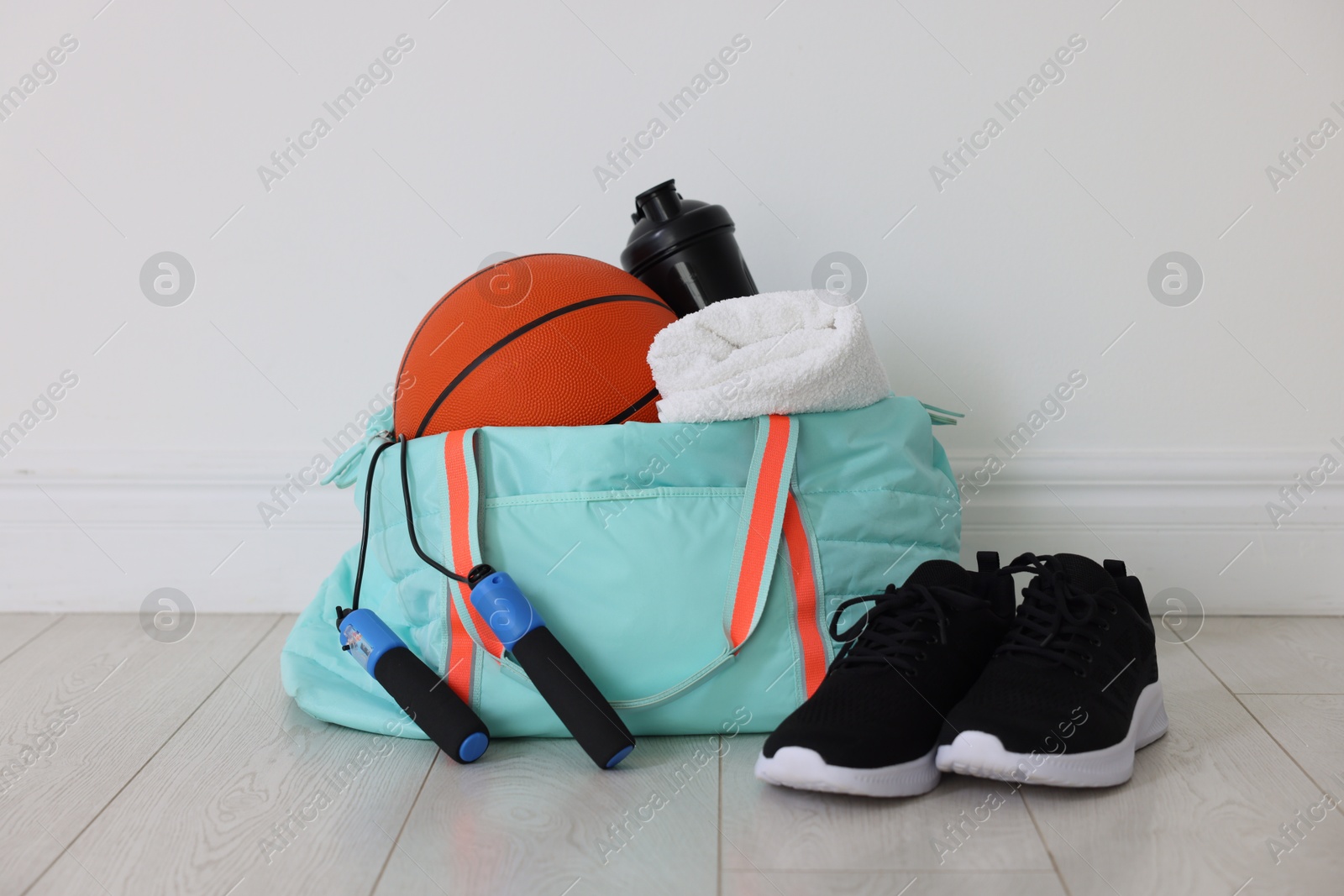 Photo of Stylish sports bag with basketball ball, skipping rope, water bottle, towel and sneakers on floor near white wall indoors