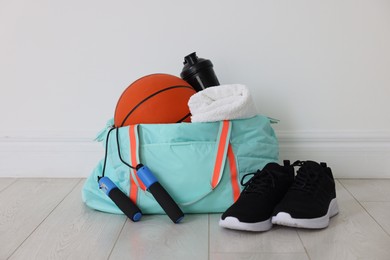 Photo of Stylish sports bag with basketball ball, skipping rope, water bottle, towel and sneakers on floor near white wall indoors