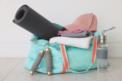 Photo of Stylish sports bag with sportswear, fitness mat, skipping rope, towel and water bottle on floor near white wall indoors