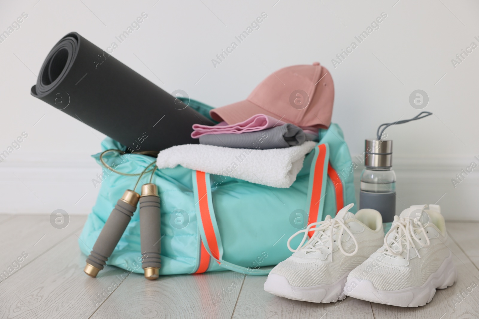 Photo of Stylish sports bag with sportswear, fitness mat, skipping rope, towel, water bottle and sneakers on floor near white wall indoors, closeup
