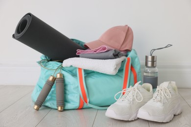 Photo of Stylish sports bag with sportswear, fitness mat, skipping rope, towel, water bottle and sneakers on floor near white wall indoors, closeup
