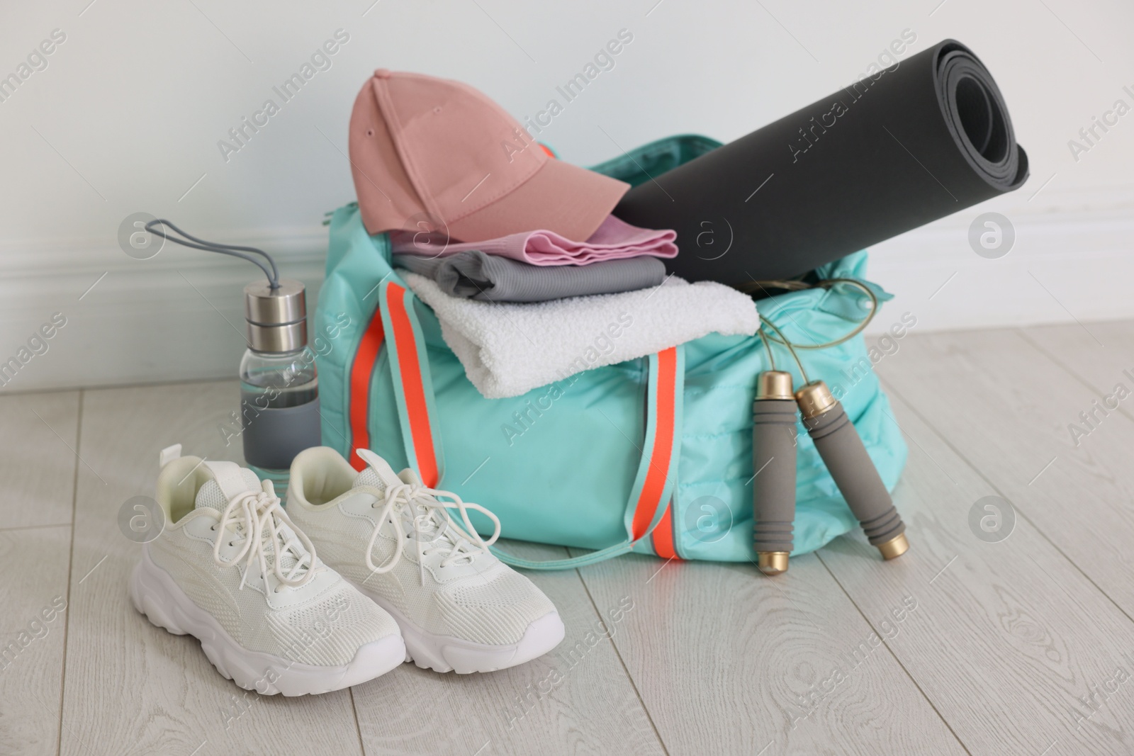 Photo of Stylish sports bag with sportswear, fitness mat, skipping rope, towel, water bottle and sneakers on floor near white wall indoors