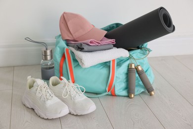 Photo of Stylish sports bag with sportswear, fitness mat, skipping rope, towel, water bottle and sneakers on floor near white wall indoors