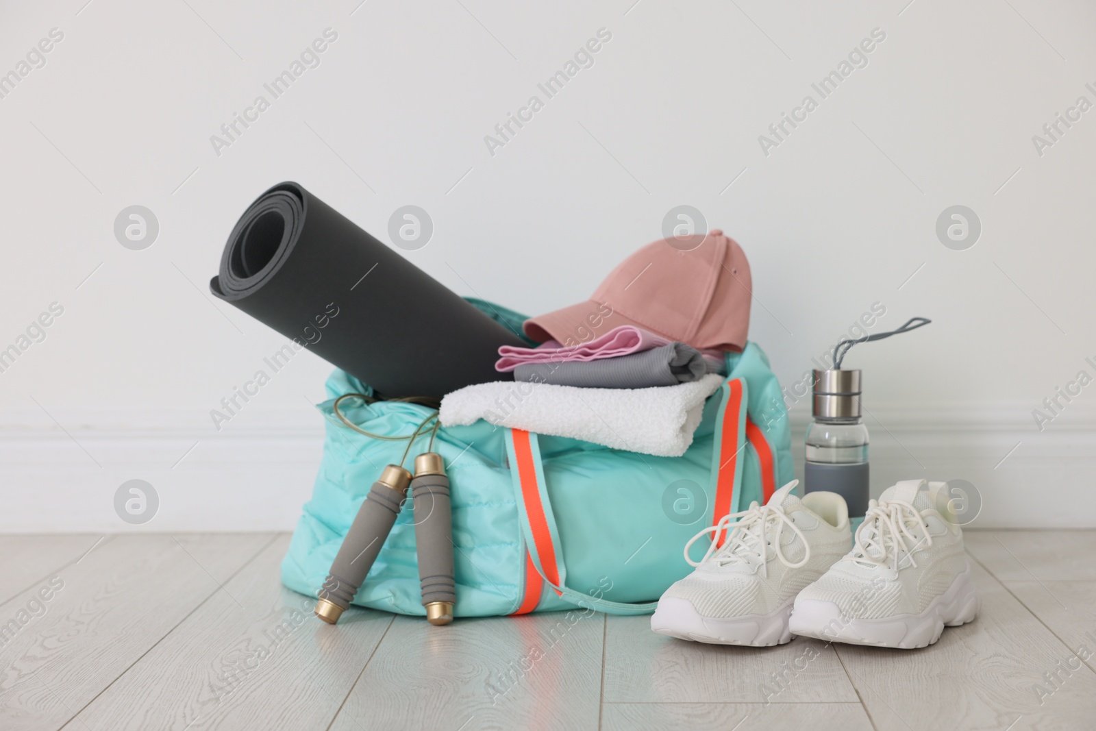 Photo of Stylish sports bag with sportswear, fitness mat, skipping rope, towel, water bottle and sneakers on floor near white wall indoors