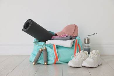 Photo of Stylish sports bag with sportswear, fitness mat, skipping rope, towel, water bottle and sneakers on floor near white wall indoors