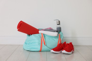 Photo of Stylish sports bag with sportswear, fitness mat, towel, water bottle and sneakers on floor near white wall indoors