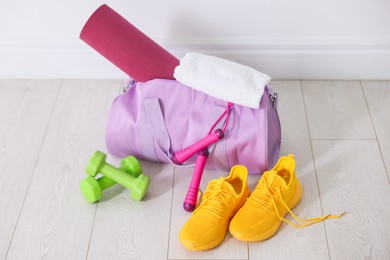 Photo of Stylish sports bag with fitness mat, towel, skipping rope, dumbbells and sneakers on floor near white wall indoors
