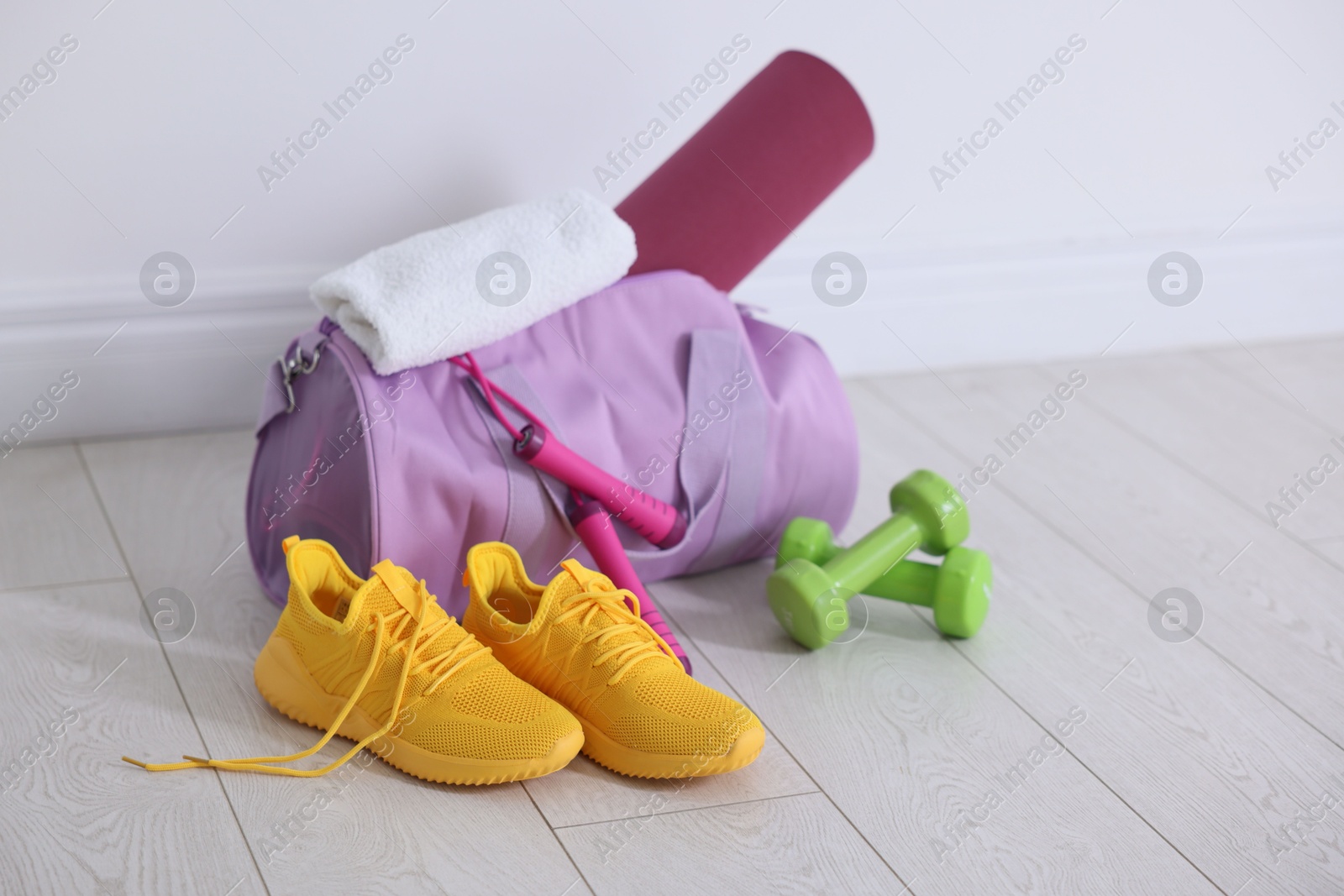 Photo of Stylish sports bag with fitness mat, towel, skipping rope, dumbbells and sneakers on floor near white wall indoors, space for text
