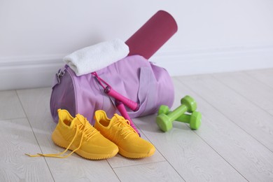 Photo of Stylish sports bag with fitness mat, towel, skipping rope, dumbbells and sneakers on floor near white wall indoors, space for text