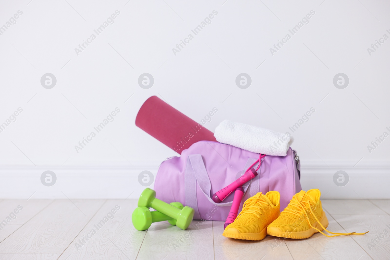 Photo of Stylish sports bag with fitness mat, towel, skipping rope, dumbbells and sneakers on floor near white wall indoors, space for text