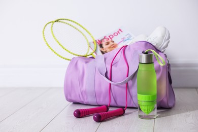Photo of Stylish sports bag with badminton rackets, magazine, sneakers, skipping rope and water bottle on floor near white wall indoors, closeup. Space for text