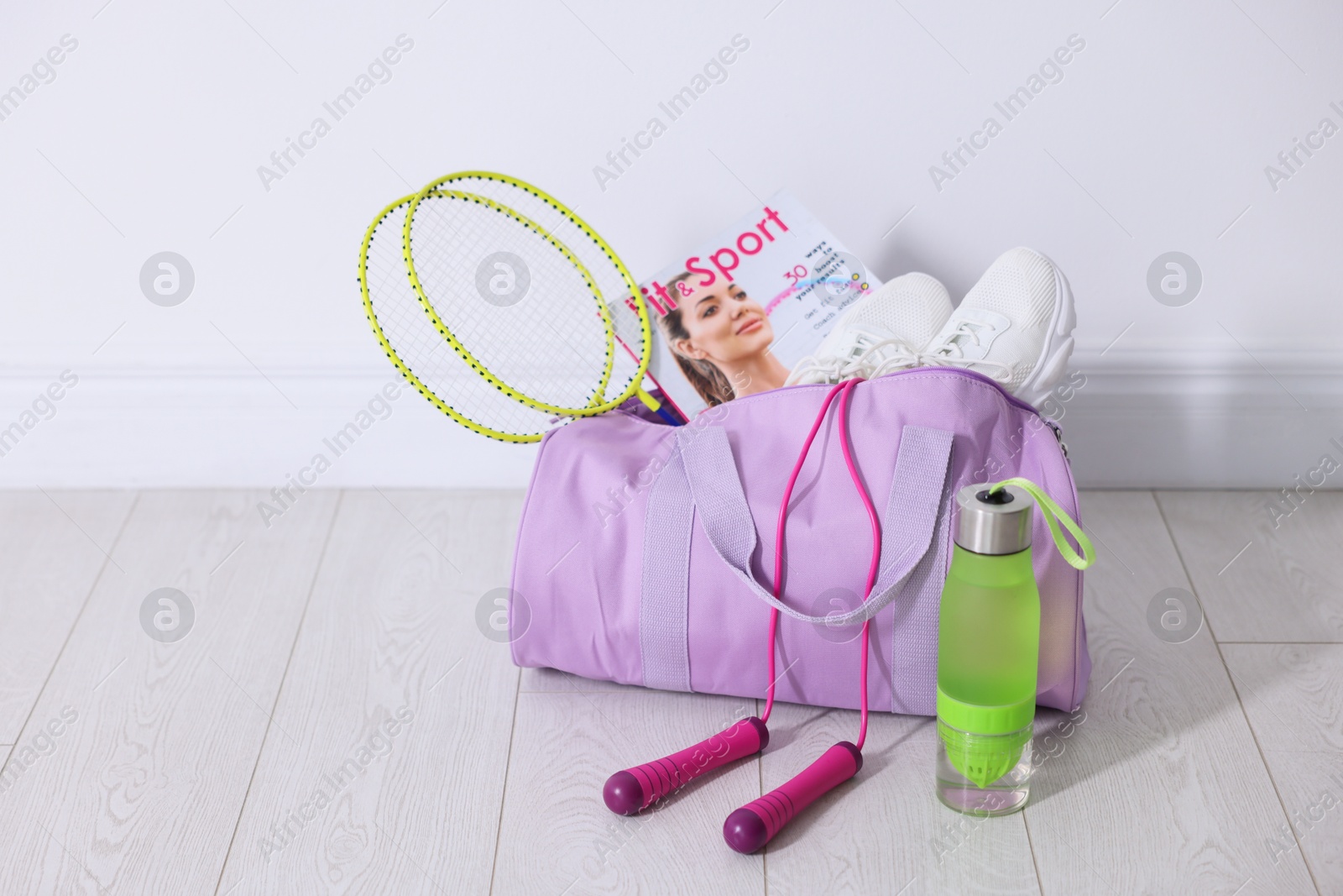 Photo of Stylish sports bag with badminton rackets, magazine, sneakers, skipping rope and water bottle on floor near white wall indoors, space for text