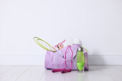 Photo of Stylish sports bag with badminton rackets, magazine, sneakers, skipping rope and water bottle on floor near white wall indoors