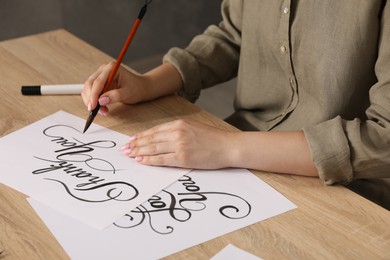 Photo of Calligraphy. Woman with brush writing words Thank You on sheet of paper at wooden table, closeup
