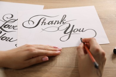 Photo of Calligraphy. Woman with brush writing words Thank You on sheet of paper at wooden table, closeup