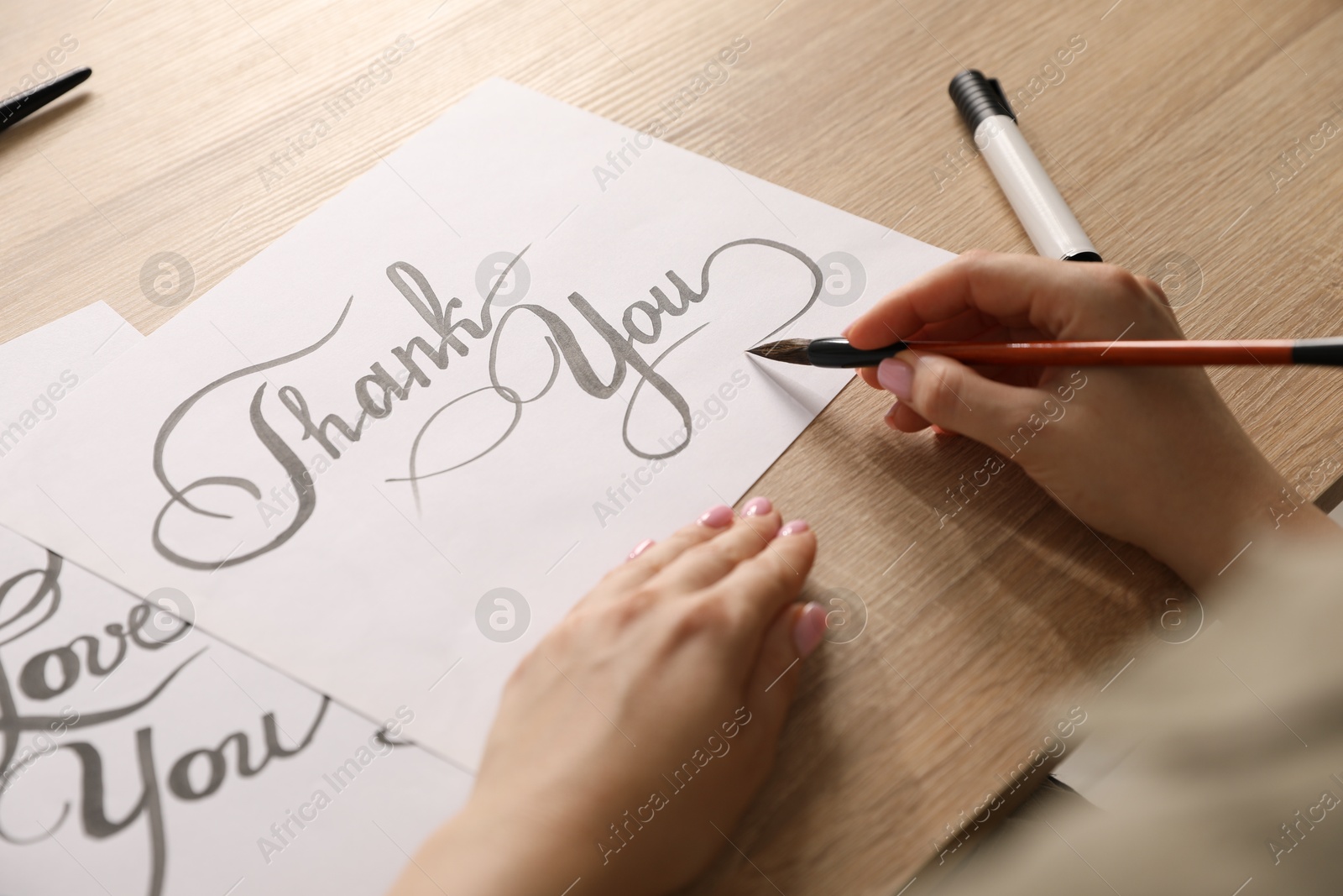 Calligraphy. Woman with brush writing words Thank You on sheet of paper at wooden table, closeup Photo of Calligraphy. Woman with brush writing words Thank You on sheet of paper at wooden table, closeup