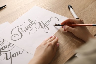 Photo of Calligraphy. Woman with brush writing words Thank You on sheet of paper at wooden table, closeup