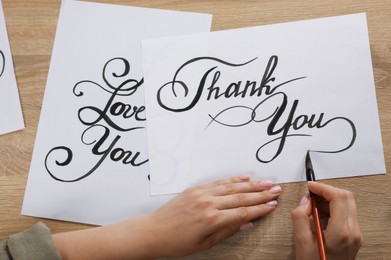 Photo of Calligraphy. Woman with brush writing words Thank You on sheet of paper at wooden table, top view