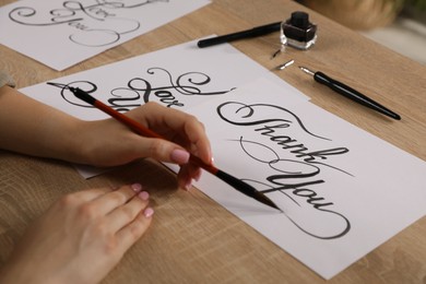 Photo of Calligraphy. Woman with brush writing words Thank You on sheet of paper at wooden table, closeup