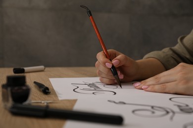 Photo of Calligraphy. Woman with brush writing words Thank You on sheet of paper at wooden table, closeup