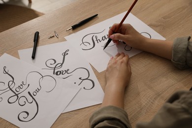 Photo of Calligraphy. Woman with brush writing words Thank You on sheet of paper at wooden table, closeup