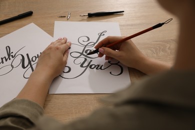 Photo of Calligraphy. Woman with brush writing phrase I Love You on sheet of paper at wooden table, closeup