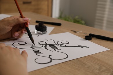 Photo of Calligraphy. Woman with brush writing phrase I Love You on sheet of paper at wooden table, closeup