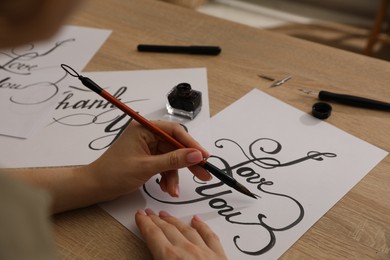 Photo of Calligraphy. Woman with brush writing phrase I Love You on sheet of paper at wooden table, closeup