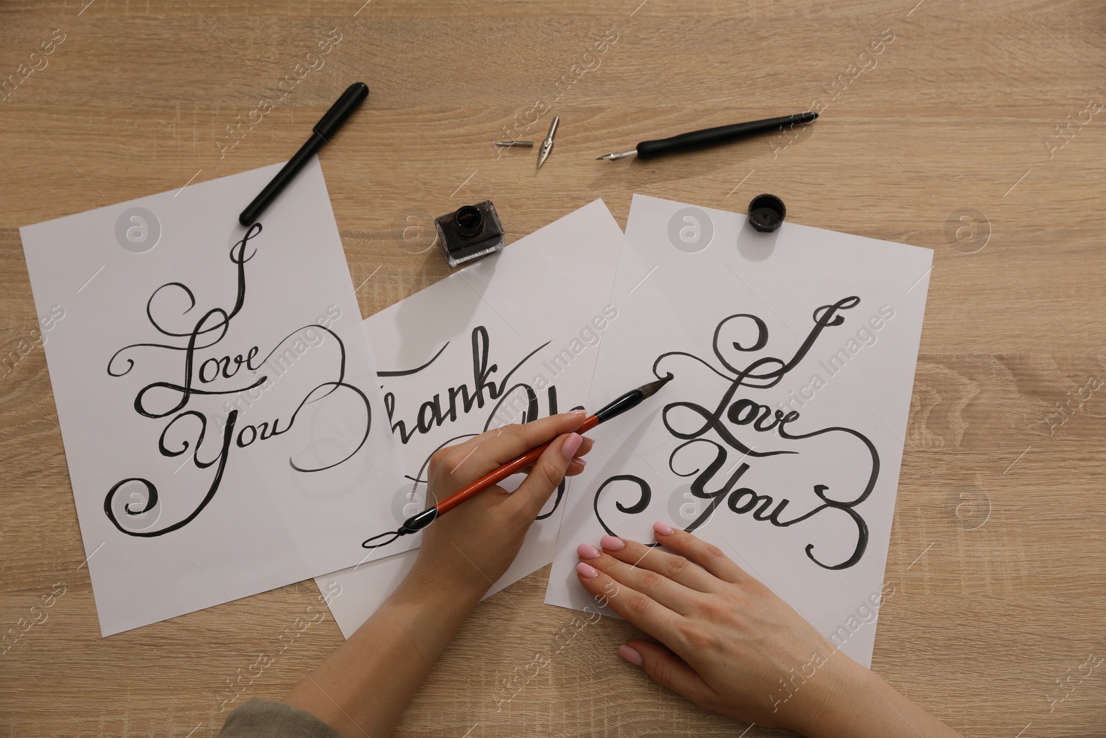 Photo of Calligraphy. Woman with brush writing phrase I Love You on sheet of paper at wooden table, top view