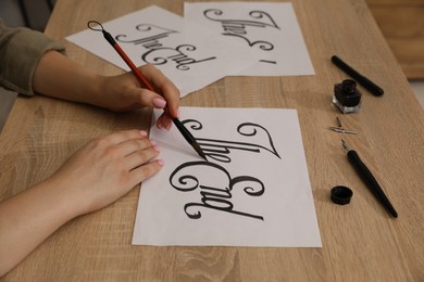 Photo of Calligraphy. Woman with brush writing words The End on sheet of paper at wooden table, closeup