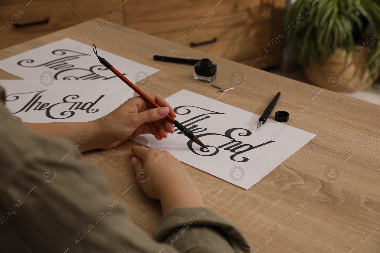 Photo of Calligraphy. Woman with brush writing words The End on sheet of paper at wooden table, closeup