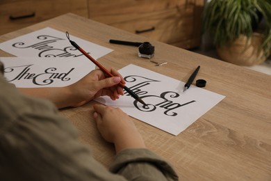 Photo of Calligraphy. Woman with brush writing words The End on sheet of paper at wooden table, closeup