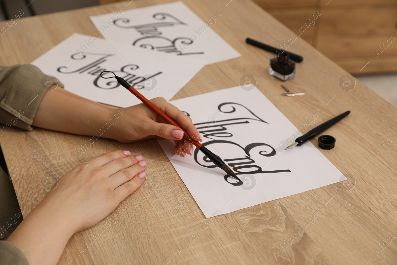 Photo of Calligraphy. Woman with brush writing words The End on sheet of paper at wooden table, closeup