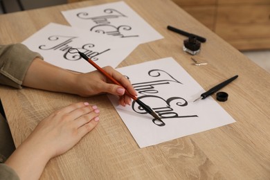 Photo of Calligraphy. Woman with brush writing words The End on sheet of paper at wooden table, closeup