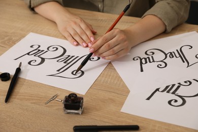 Photo of Calligraphy. Woman with brush writing words The End on sheet of paper at wooden table, closeup