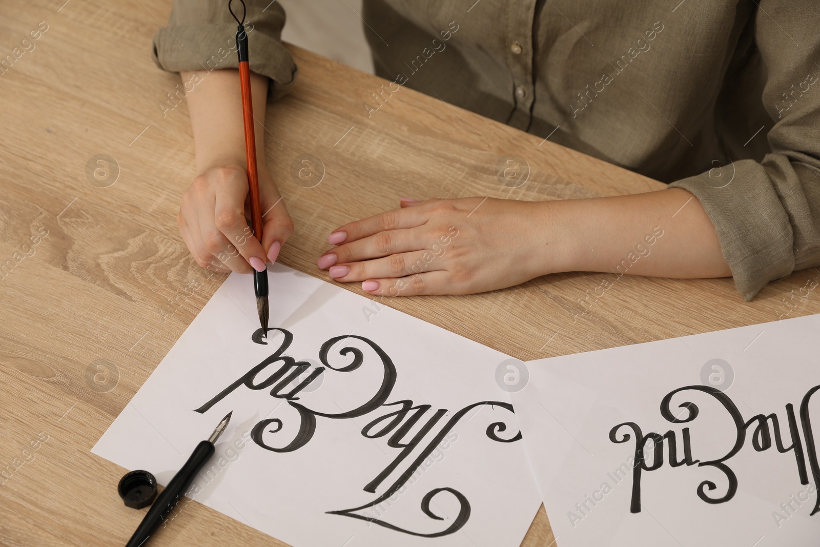 Photo of Calligraphy. Woman with brush writing words The End on sheet of paper at wooden table, closeup