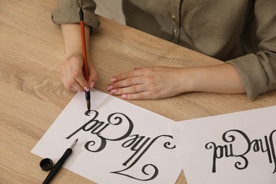 Photo of Calligraphy. Woman with brush writing words The End on sheet of paper at wooden table, closeup