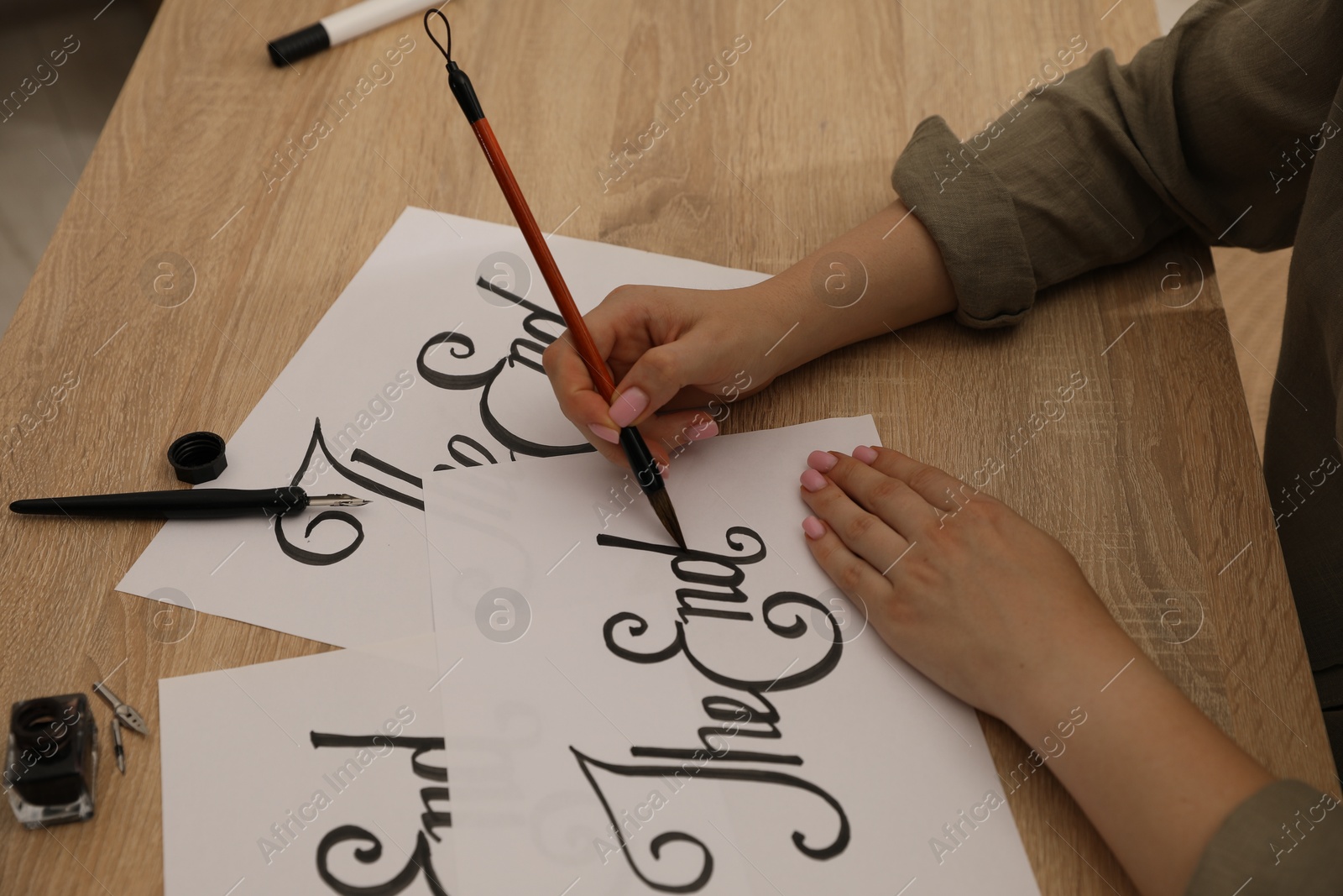 Photo of Calligraphy. Woman with brush writing words The End on sheet of paper at wooden table, closeup