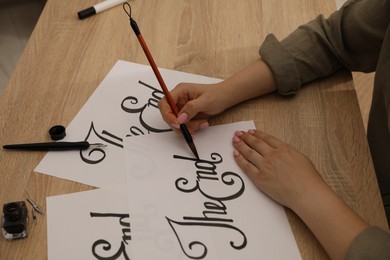 Photo of Calligraphy. Woman with brush writing words The End on sheet of paper at wooden table, closeup