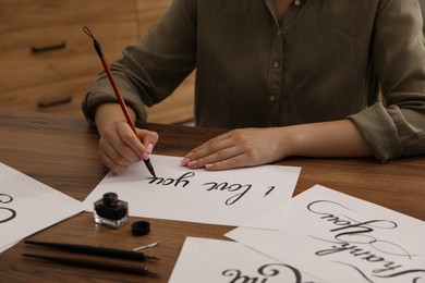 Photo of Calligraphy. Woman with brush writing phrase I Love You on sheet of paper at wooden table, closeup