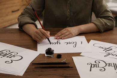 Photo of Calligraphy. Woman with brush writing phrase I Love You on sheet of paper at wooden table, closeup