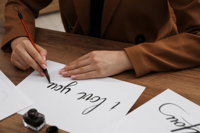 Photo of Calligraphy. Woman with brush writing phrase I Love You on sheet of paper at wooden table, closeup